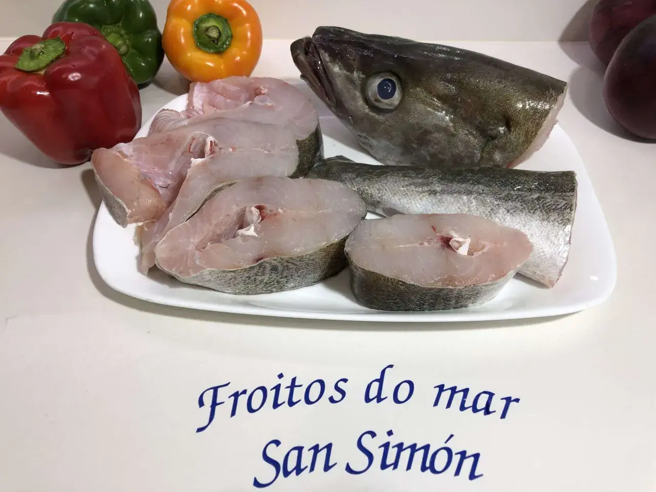 Rodajas de abadejo pescado fresco en plato blanco, junto a pimientos y el texto "Froitios do mar San Simón".