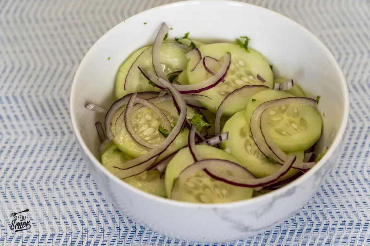 Deliciosa ensalada de pepino con cebolla morada y hierbas frescas, servida en un tazón blanco sobre un mantel azul y blanco.