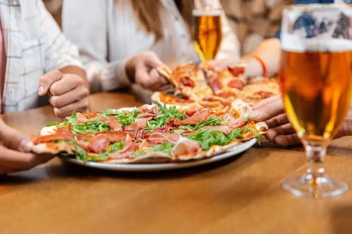 Un gruppo di amici condivide una deliziosa pizza con prosciutto e rucola, accompagnata da una fresca birra.