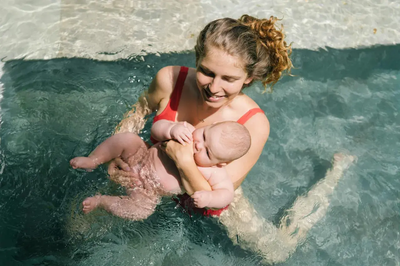 Mamma sorridente tiene in braccio il suo neonato durante un corso di nuoto per neonati in piscina.