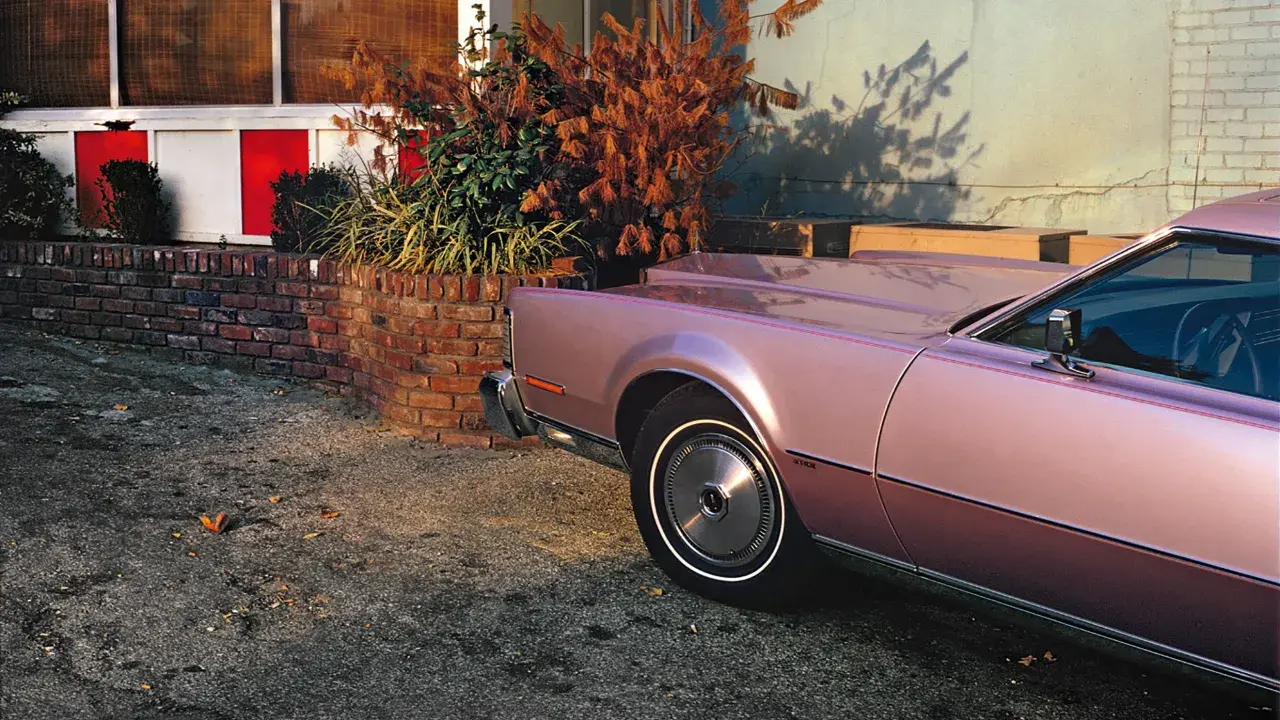 Un'iconica william eggleston foto: una Lincoln rosa metallizzato parcheggiata accanto a un muro di mattoni e un cespuglio autunnale.