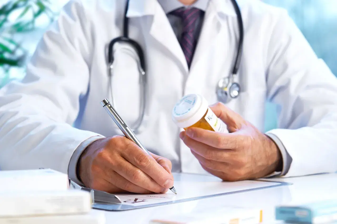 Medico con camice bianco e stetoscopio, scrive su ricetta medica, tenendo in mano un flacone di pillole. Un piano terapeutico medico di base in corso.