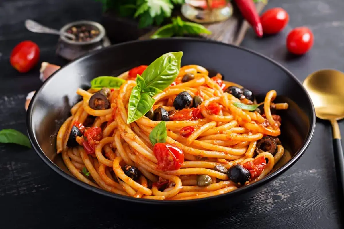 Un plat de pâtes aux anchois, tomates, olives et câpres, garni de basilic frais. Un délice méditerranéen.