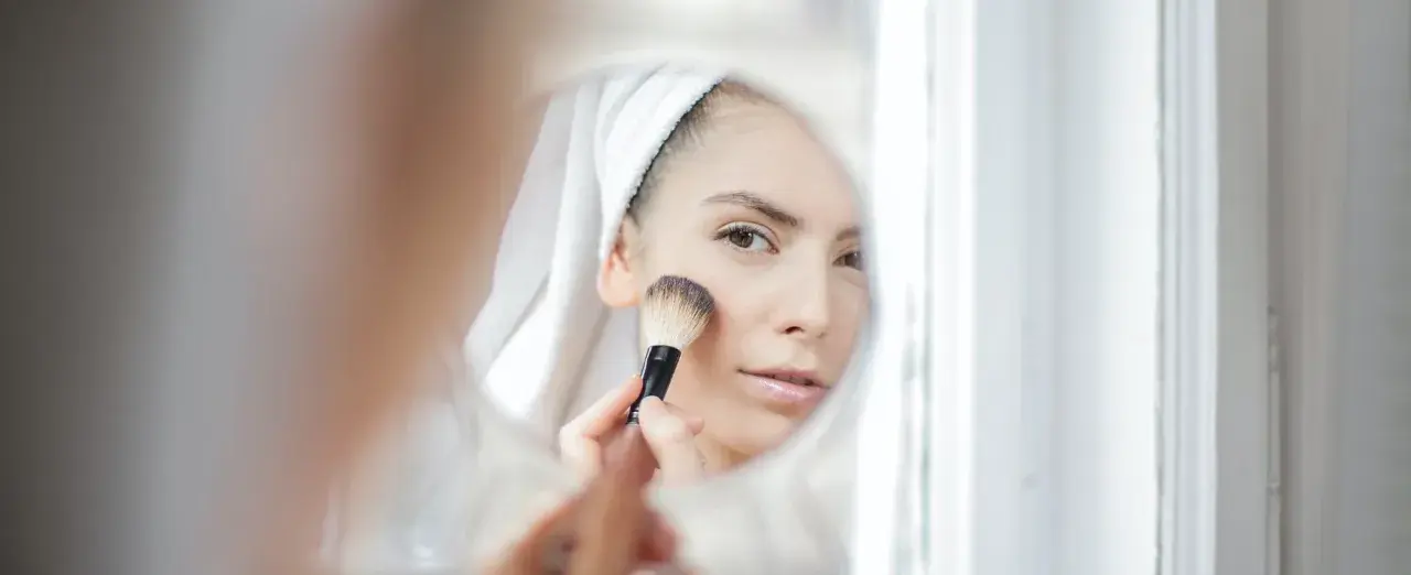 Kobieta w ręczniku na głowie nakłada puder pędzlem, patrząc w lustro. Dowiedz się, jak dobrać odcień loreal true match dla siebie.