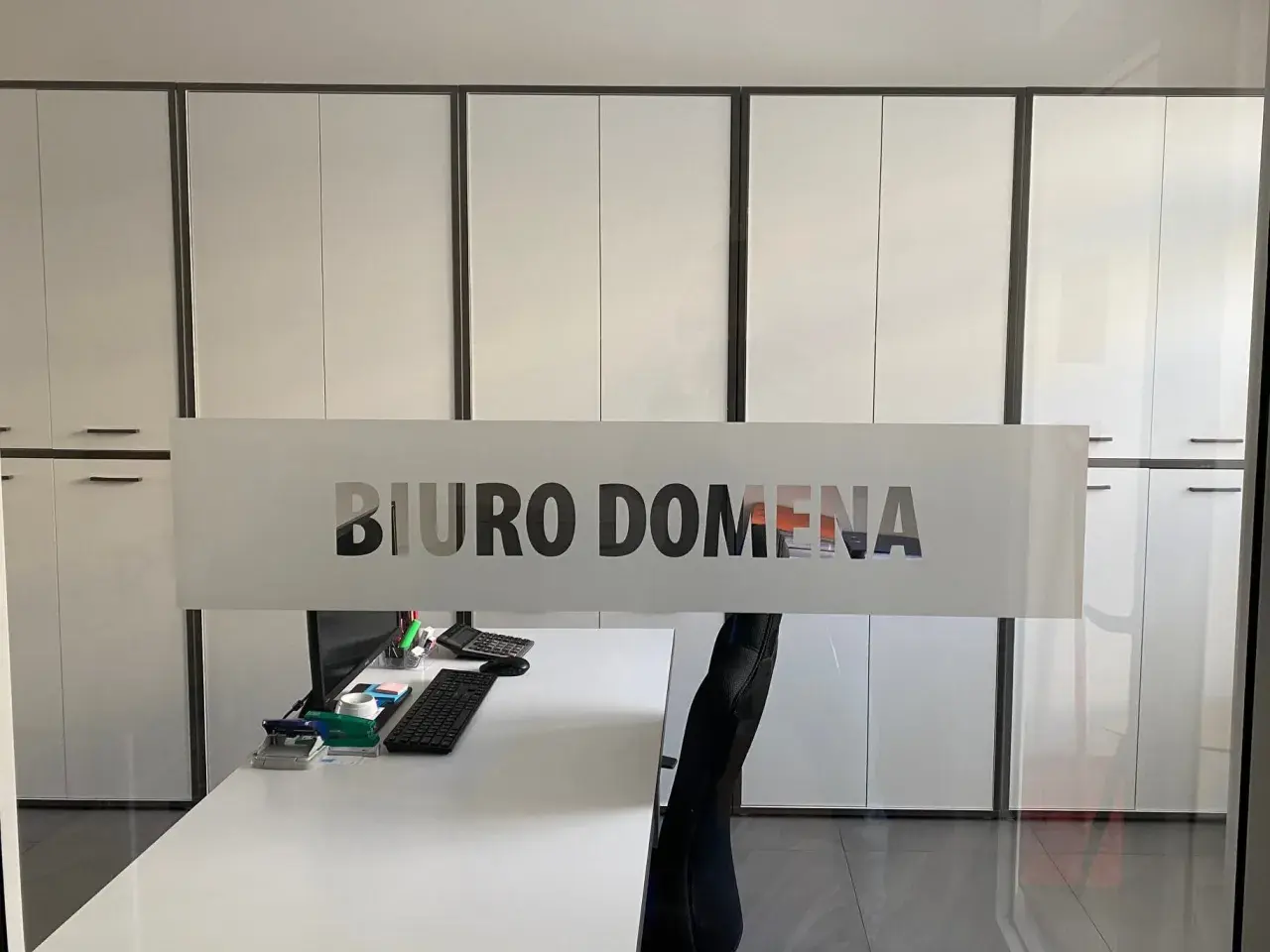 Szklana witryna z napisem "BIURO DOMENA" na tle nowoczesnego biura z białymi szafami.