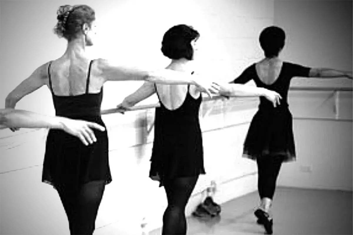 Three women in black leotards practice ballet at a barre.