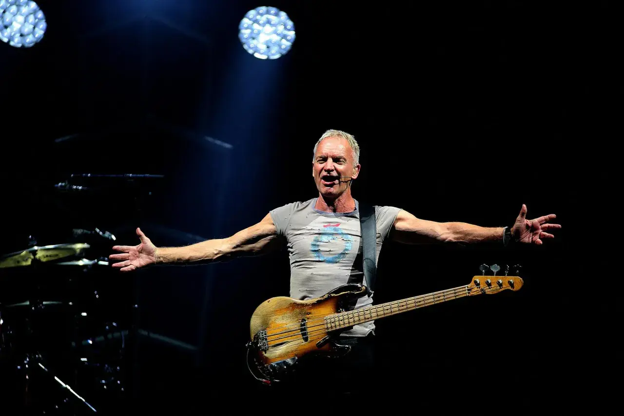 Sting śpiewa na koncercie, trzymając gitarę basową. Jego ramiona są rozłożone, jakby chciał objąć cały świat. Tekst "sting shape of my heart" idealnie pasuje do tej sceny.