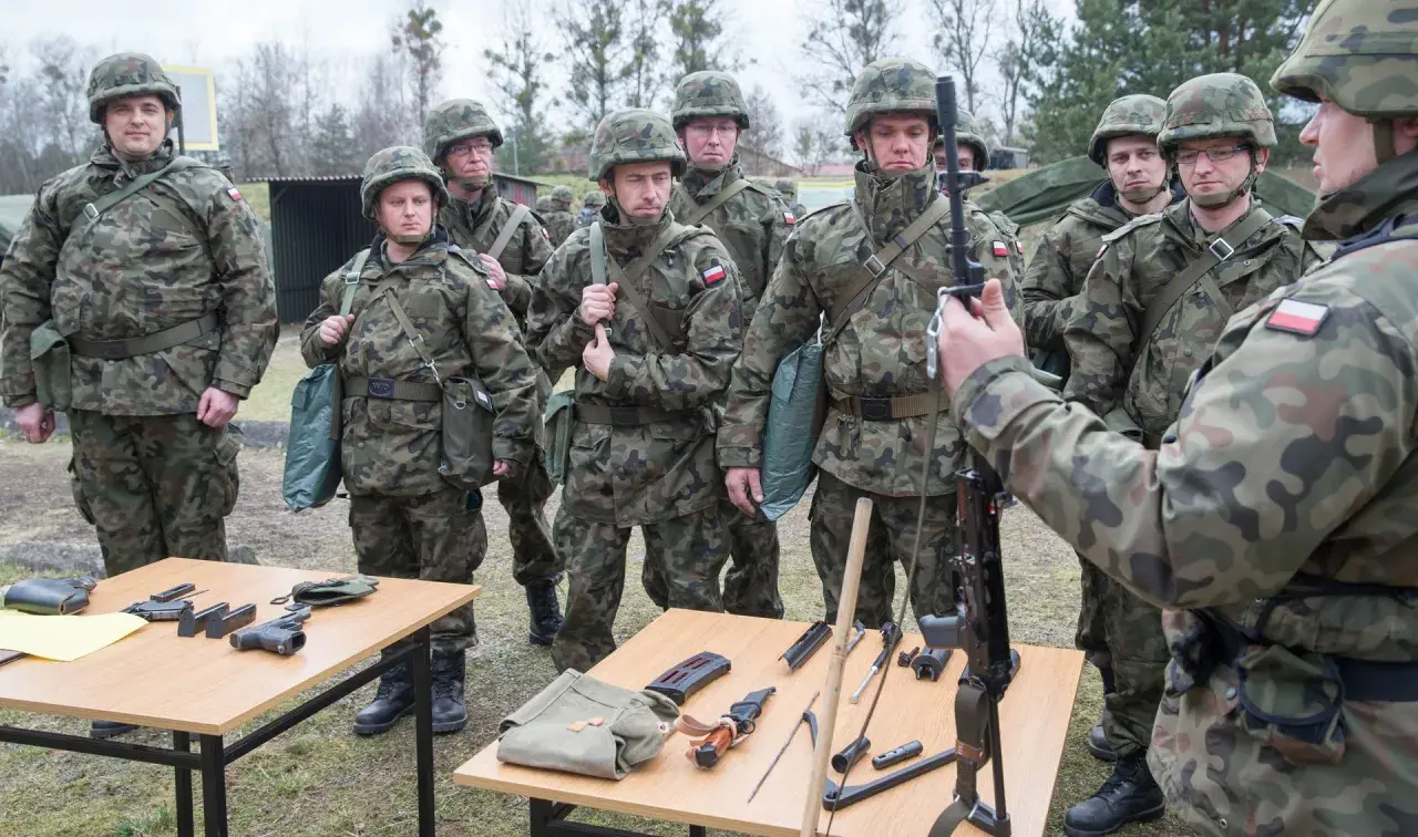 Żołnierze w mundurach ćwiczą, co zabrać na ćwiczenia rezerwy. Na stołach broń, magazynki, nóż i torby.