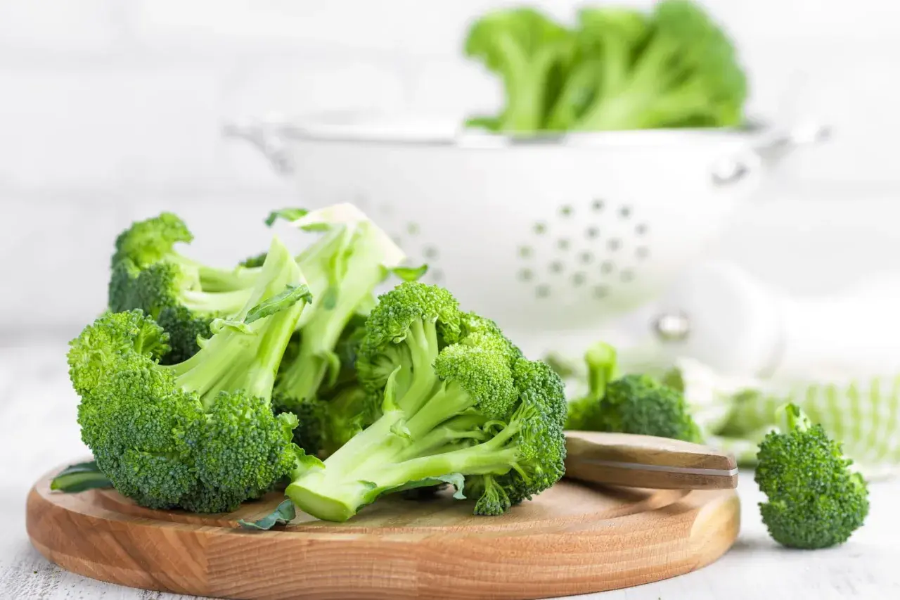 Brocolis frais coupés sur une planche en bois, prêts pour une recette saine. Un égouttoir blanc est visible en arrière-plan.