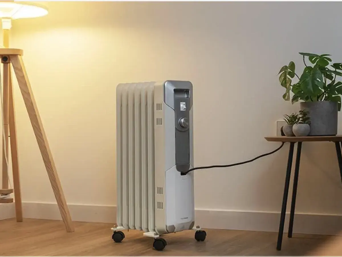 Estufa eléctrica de aceite con ruedas, conectada a la corriente, junto a una planta y una lámpara. Controla el consumo.