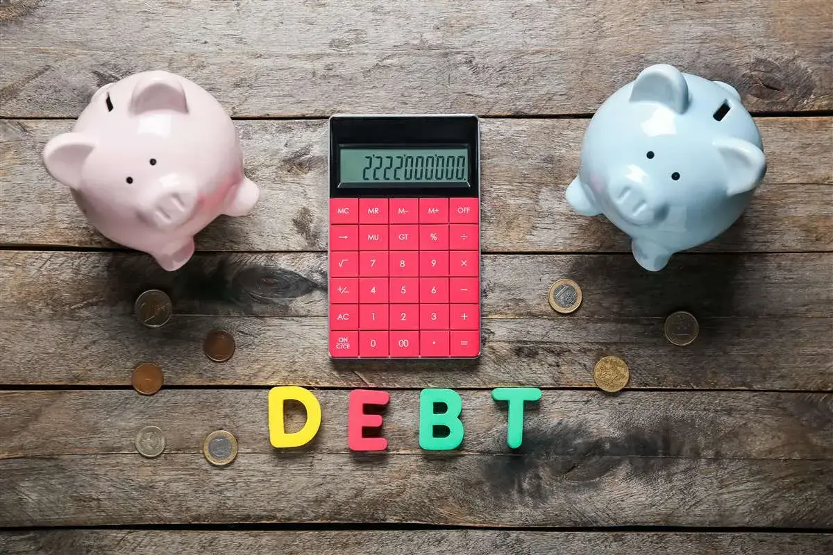 Due salvadanai rosa e azzurro, una calcolatrice rossa con un numero elevato e lettere che formano la parola "DEBT" suggeriscono un **debito residuo** da gestire.