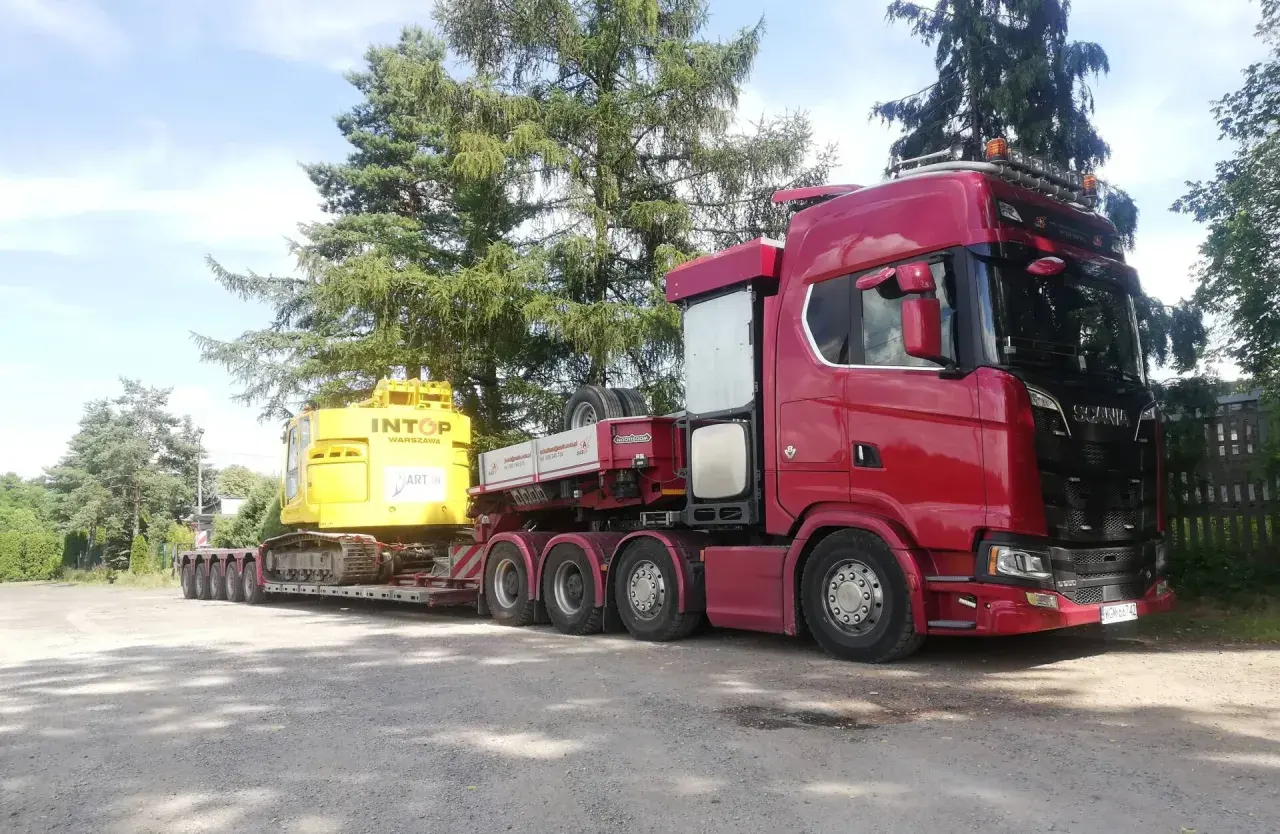 Czerwony Scania z naczepą transportuje żółtą koparkę. To typowa przesyłka dużych gabarytów, gotowa do drogi.