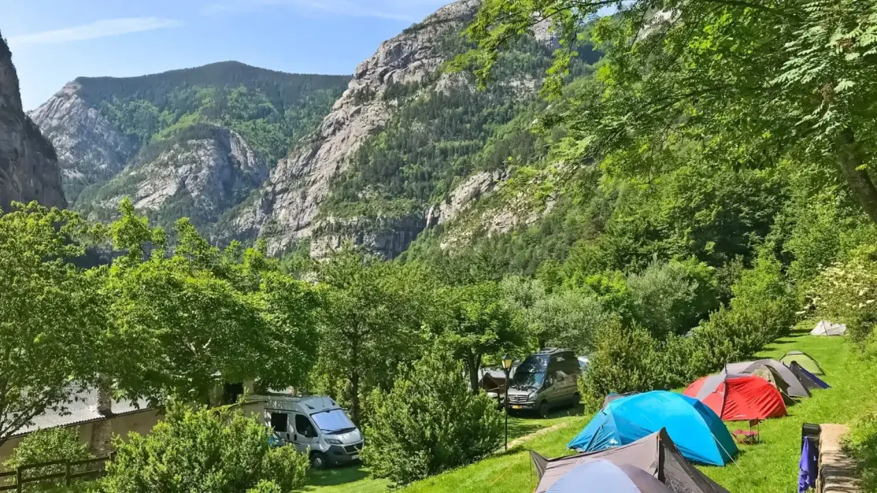 Acampar en este valle es una experiencia de los mejores camping. Tiendas de campaña y furgonetas aparcadas en un prado verde, con imponentes montañas boscosas de fondo.