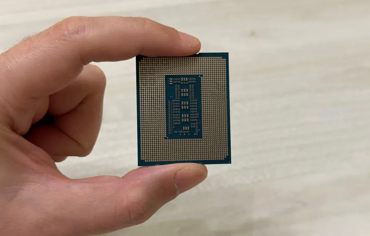 Un dedo sostiene un procesador o CPU, mostrando su parte inferior con pines dorados y un diseño rectangular en el centro.