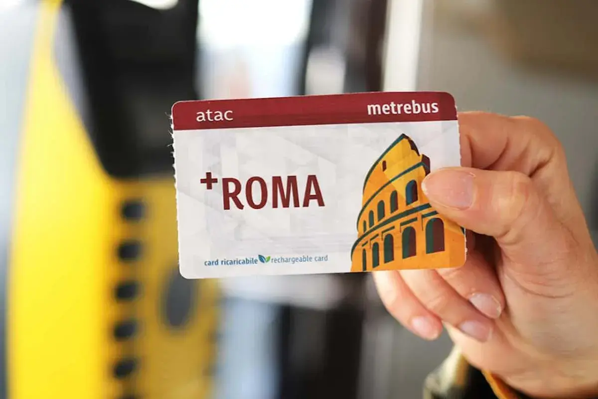 Una mano tiene una tessera metropolitana Roma con il Colosseo stilizzato. La carta è ricaricabile e riporta il logo ATAC.