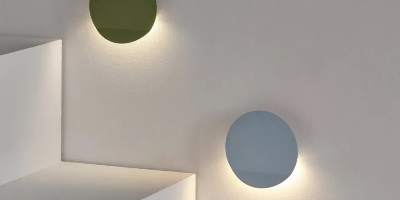 Dos lámparas de aluminio circulares, una verde y otra azul, iluminan una pared texturizada.