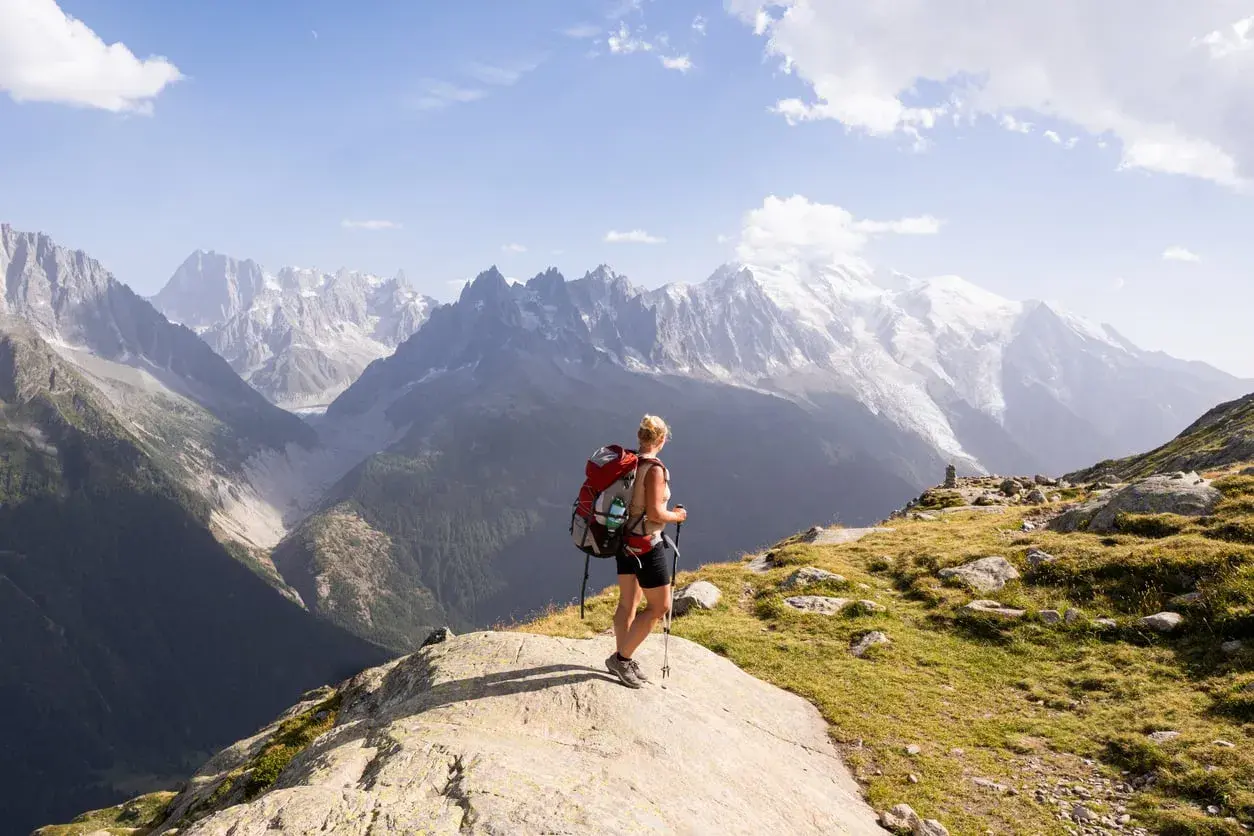 A hiker with a red backpack pauses on a rocky outcrop, enjoying the breathtaking view of the Mont Blanc massif during a challenging hike.