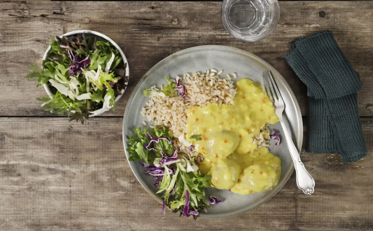 En tallrik med ris, grönsaker och krämiga boller i karry. En skål med sallad står bredvid.