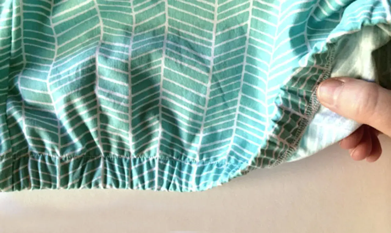 Close-up of a hand demonstrating how to sew in elastic on a teal and white chevron patterned fabric. The elastic creates a gathered, ruffled effect.