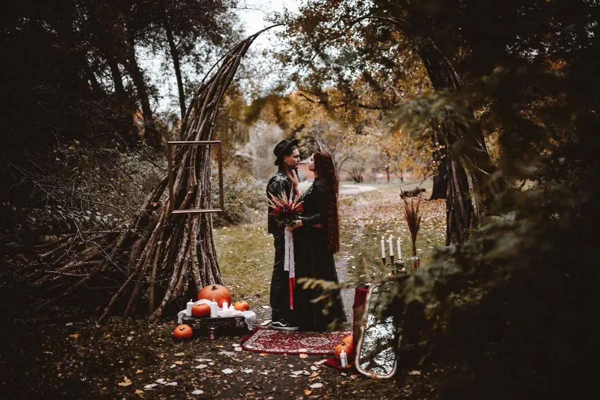 Para w czarnych strojach pod łukiem z gałęzi, dynie i świece.