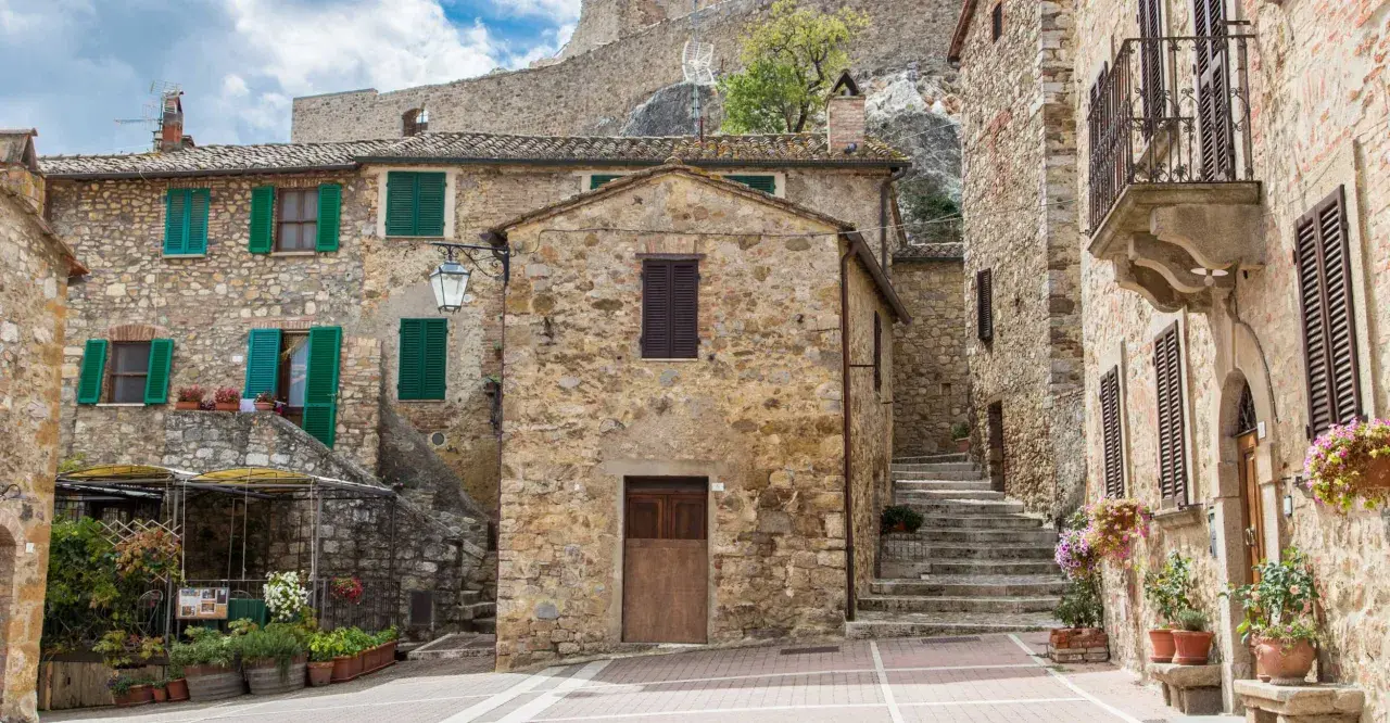 Un vicolo pittoresco a Castel d'Orcia, con case in pietra, persiane verdi e fiori colorati.