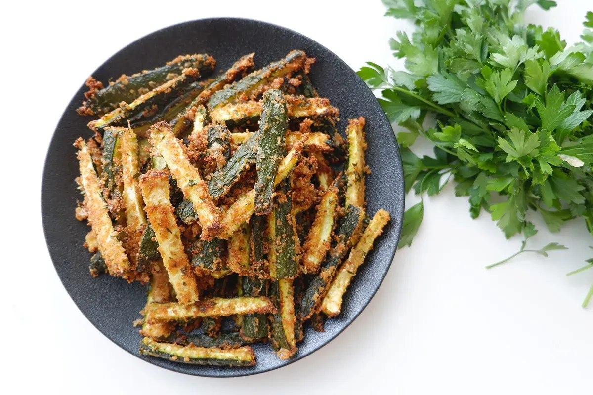 Zucchine gratinate in padella, dorate e croccanti, servite su un piatto nero con prezzemolo fresco.