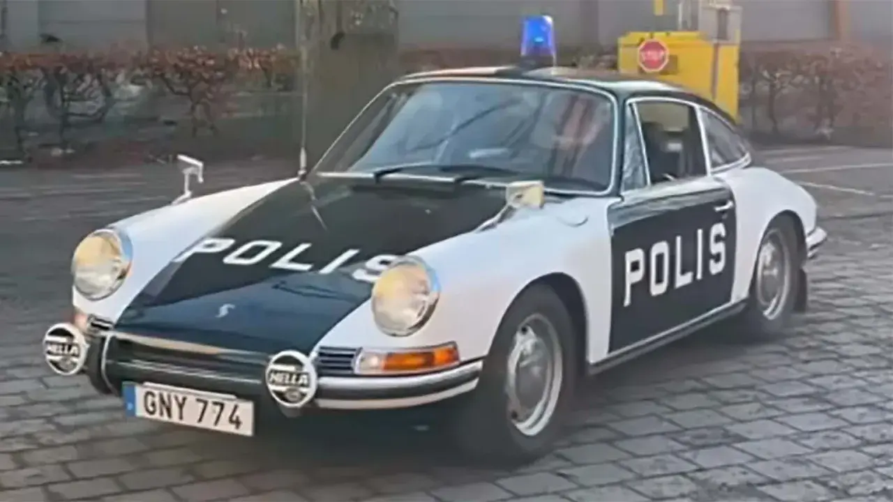 En klassisk Porsche polisbil med blåljus på taket. Bilen är vit med svarta detaljer och texten "POLIS" på motorhuven och dörren.