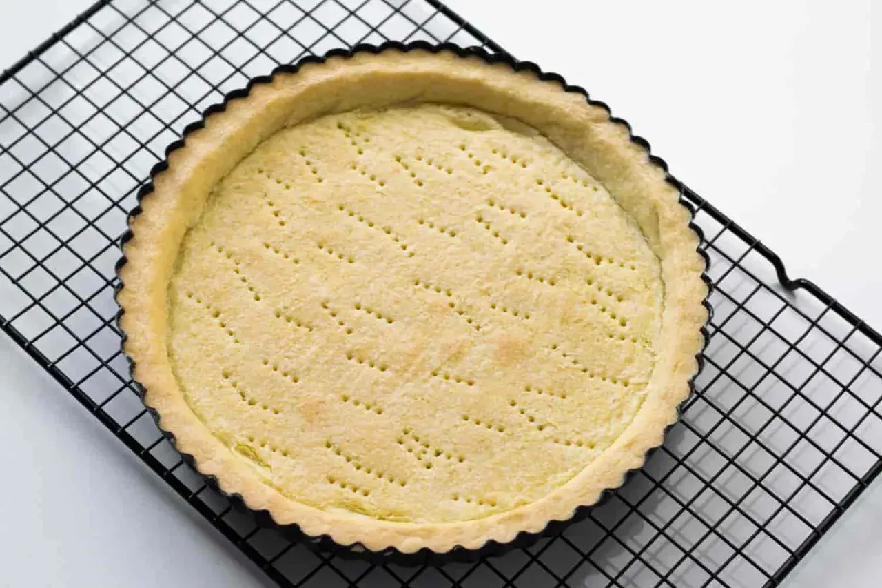 Base di pasta sucrée dorata e forata, pronta per essere farcita, su una griglia nera.