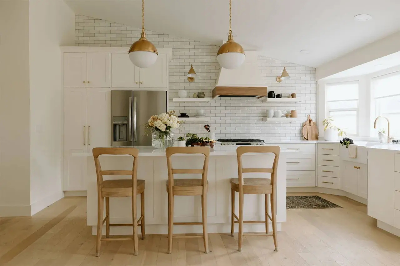 A bright, airy modern farmhouse kitchen with white subway tile, gold pendant lights, and a large island with three wooden stools.