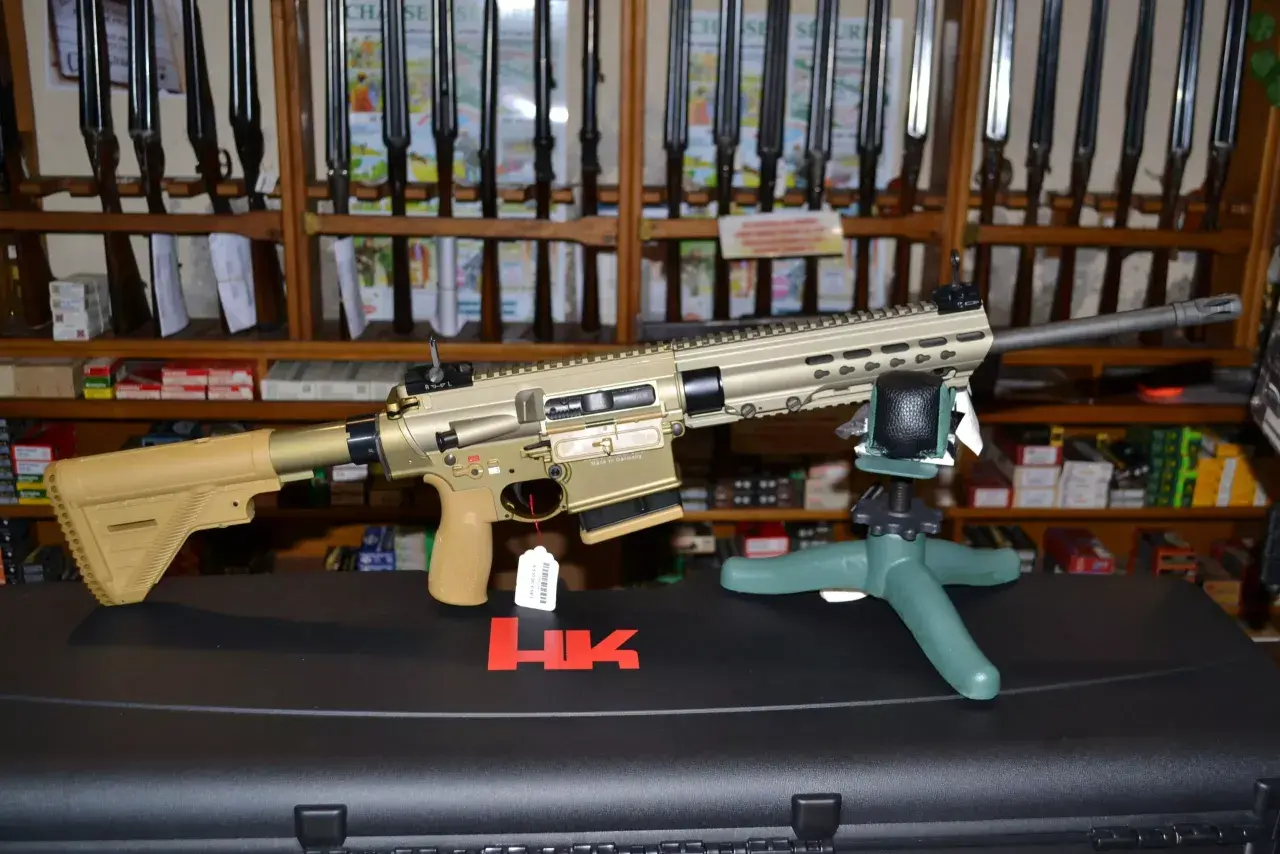 Un fusil HK MR 308, de couleur sable, est présenté sur un support. Derrière, des étagères chargées de fusils et de boîtes de munitions.