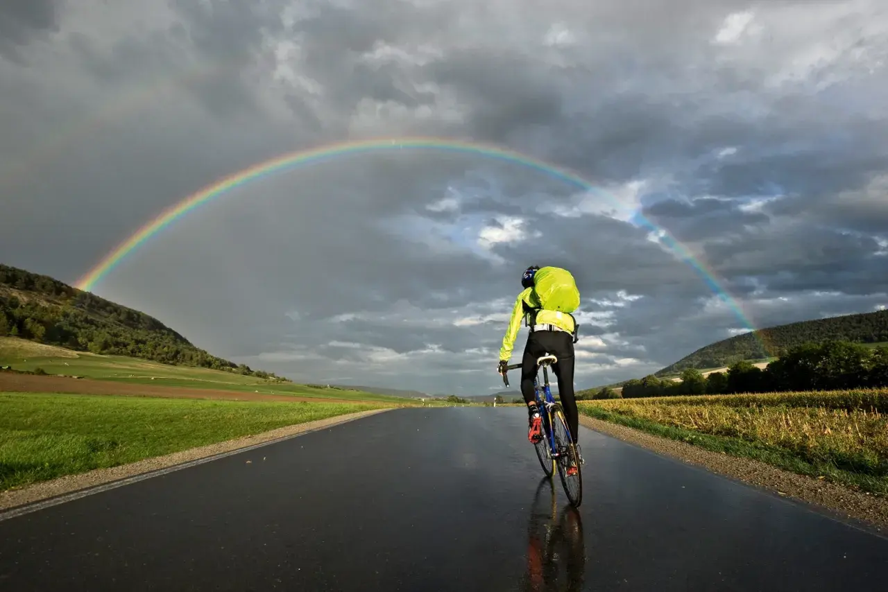 Rennradtour 3 Tage: Ein Radfahrer fährt unter einem Regenbogen auf nasser Straße.