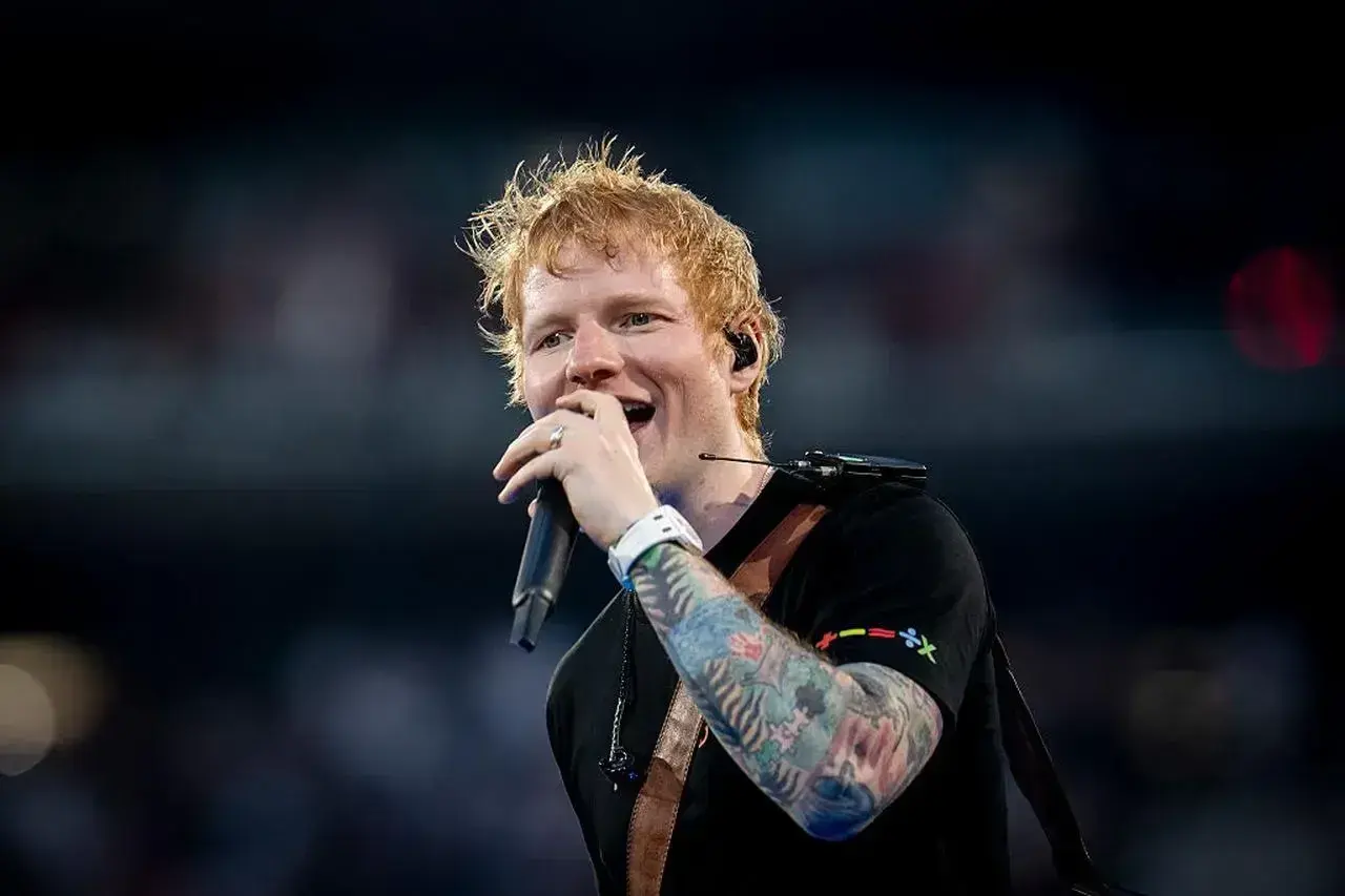 Ed Sheeran śpiewa z mikrofonem, jego ręka ozdobiona kolorowymi tatuażami.