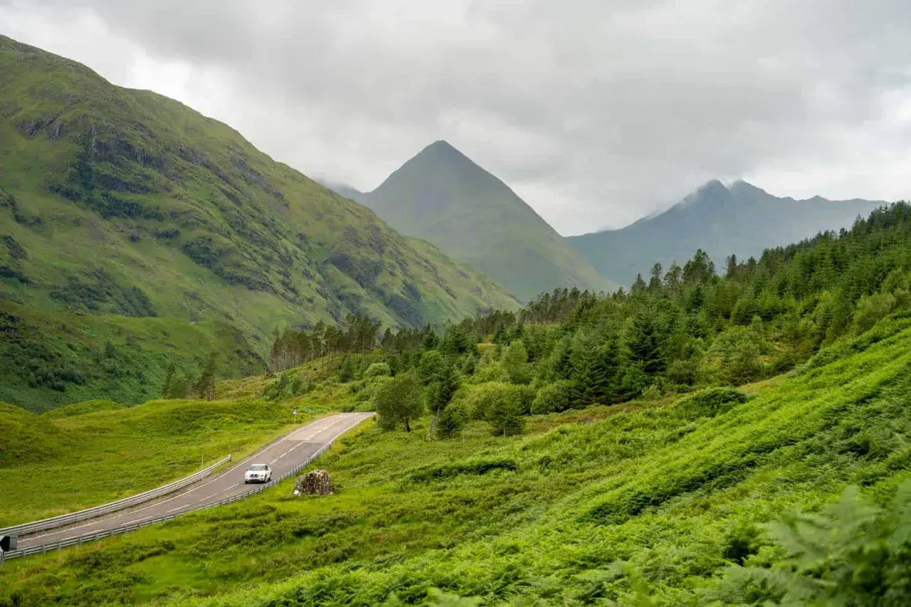 A car drives on a winding road through lush green Scottish Highlands, perfect for a hiking itinerary.