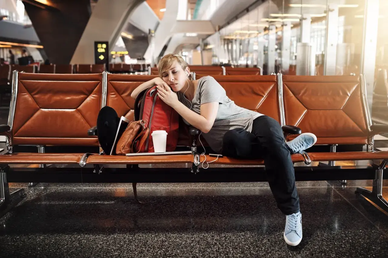Zmęczona podróżniczka śpi na lotnisku, próbując pokonać jetlag. Obok niej plecak i kubek kawy.