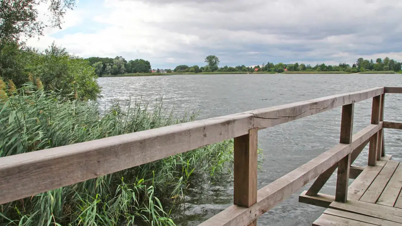 Drewniany pomost nad jeziorem, gdzie można łowić ryby. W oddali widać zalesiony brzeg i domy.