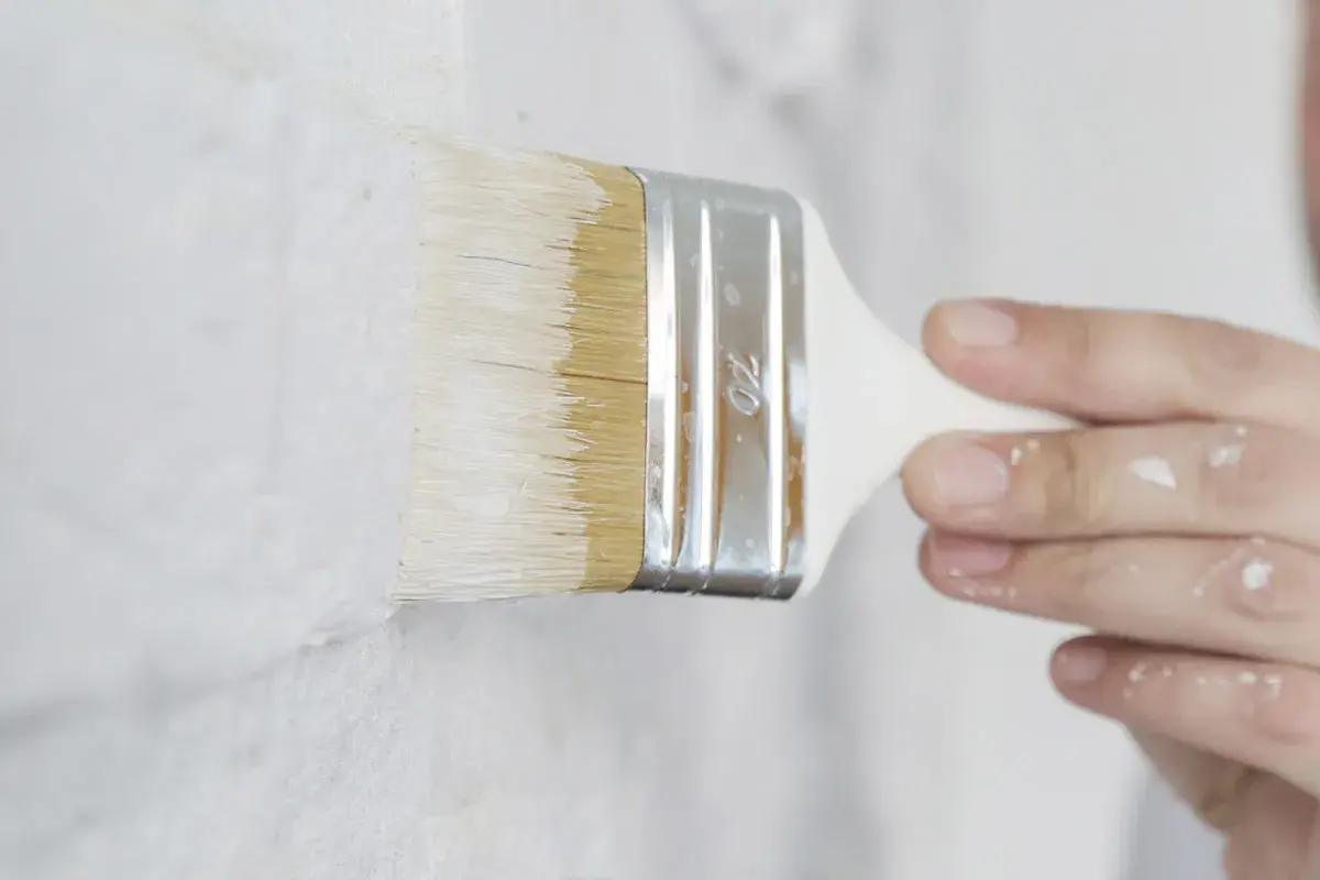 Pennello bianco stende pittura su muro grezzo. Si può mischiare il fissativo nella pittura per una finitura migliore.