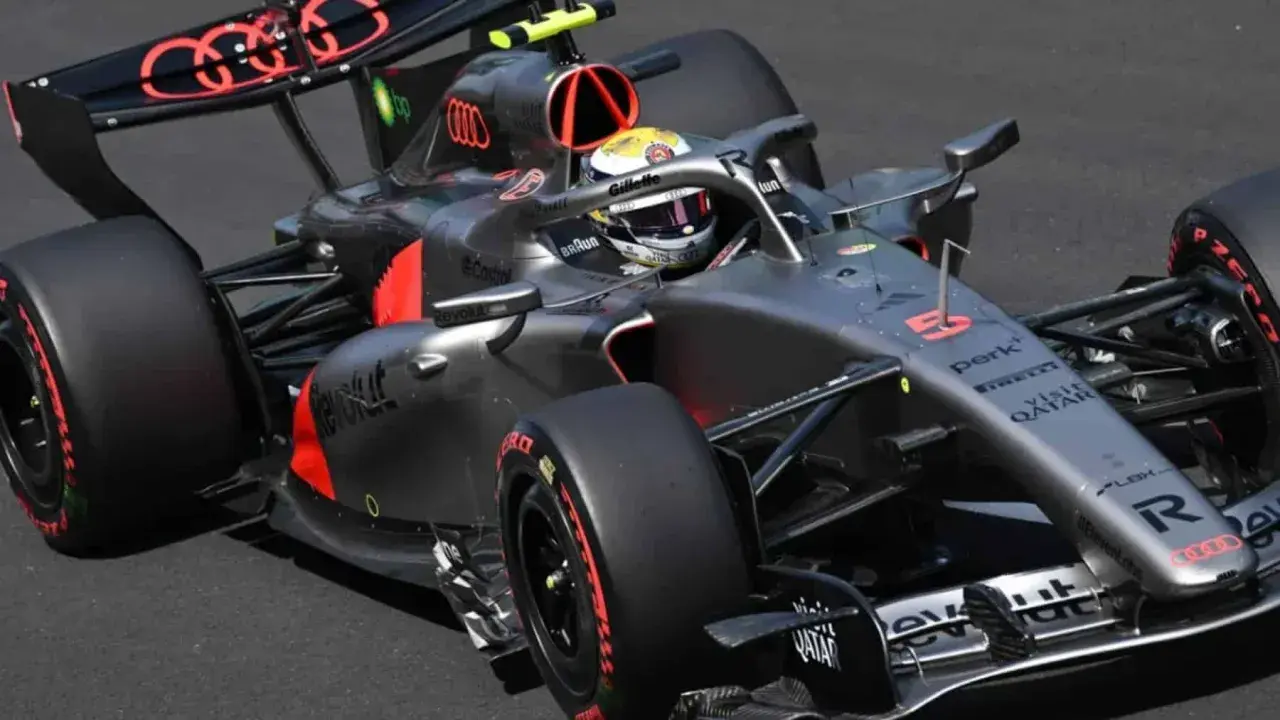 Un coche de **Audi F1** gris y negro con detalles rojos acelera en la pista. El piloto lleva casco amarillo y blanco.