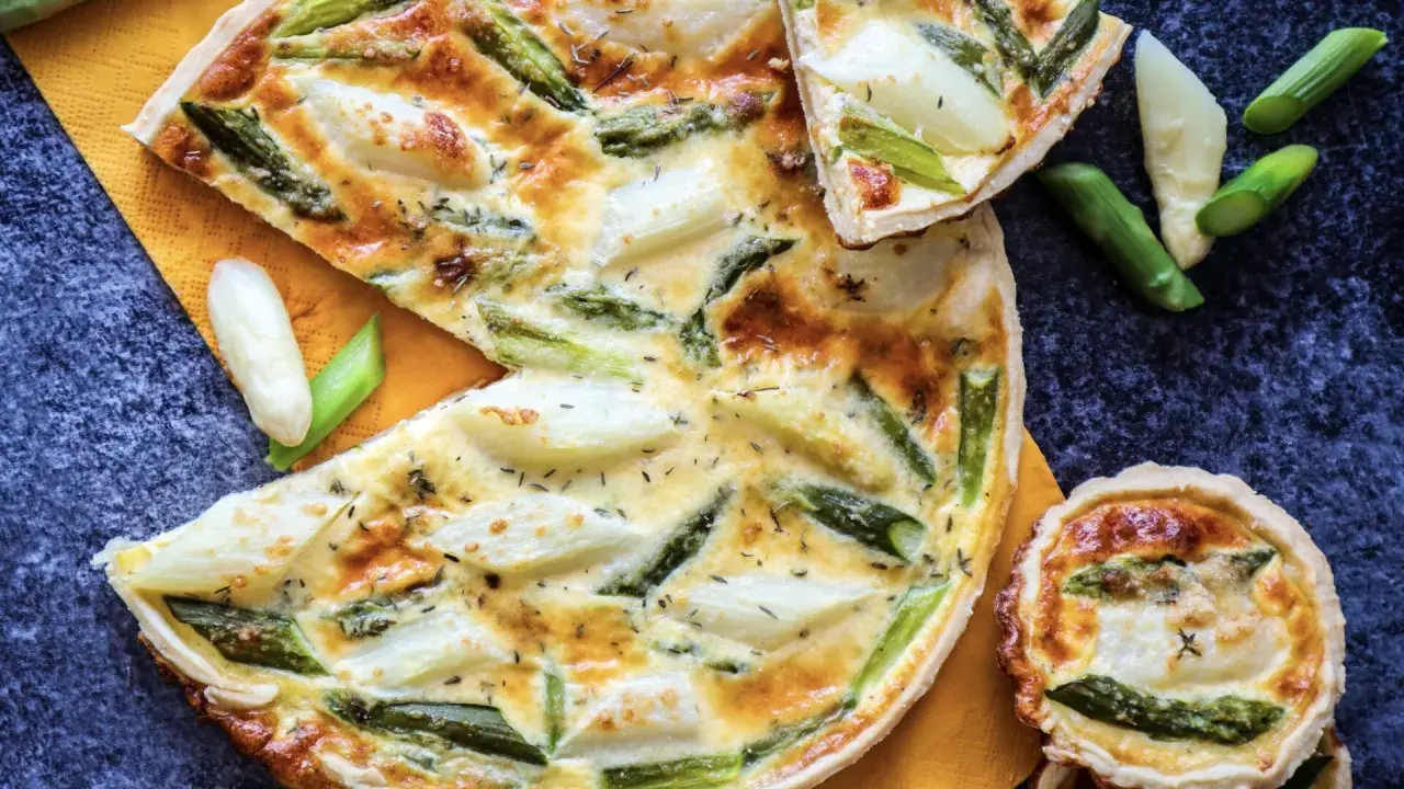 Herzhafte Quiche mit Spargel, goldbraun gebacken, auf einem orangen Tuch. Ein Stück ist angeschnitten, daneben liegen frische Spargelstücke.