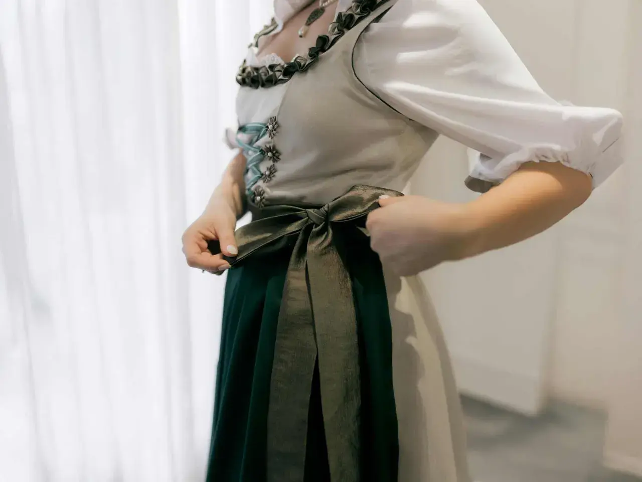 Eine Frau bindet die Schleife ihres Dirndls. Das Dirndl hat eine helle Weste mit Schnürung und eine dunkelgrüne Schürze.