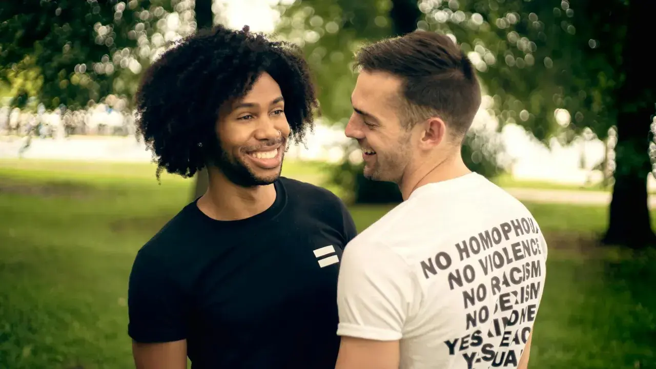 Due uomini sorridono, uno con maglia nera con simbolo di uguaglianza, l'altro con maglia bianca con messaggi di inclusione. Un'immagine che evoca il poliamore significato di amore libero e rispetto.