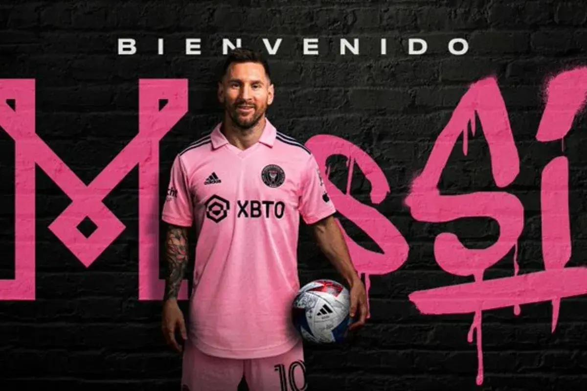 Lionel Messi w różowej koszulce Inter Miami, z piłką w ręku, na tle napisu "Bienvenido Messi". Nowy inter miami trener.