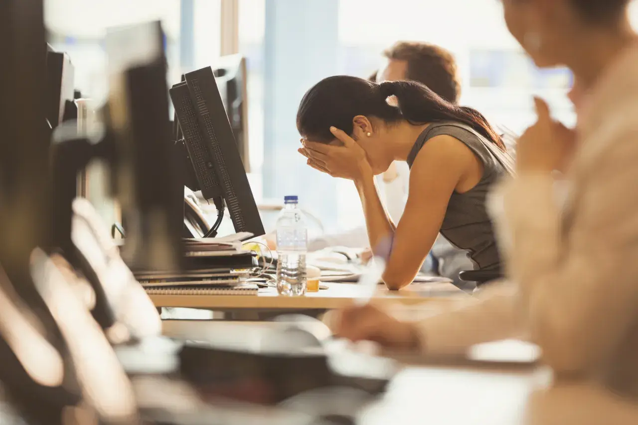 A woman sits at her desk, head in hands, looking demoralized at work. A bottle of water sits nearby.