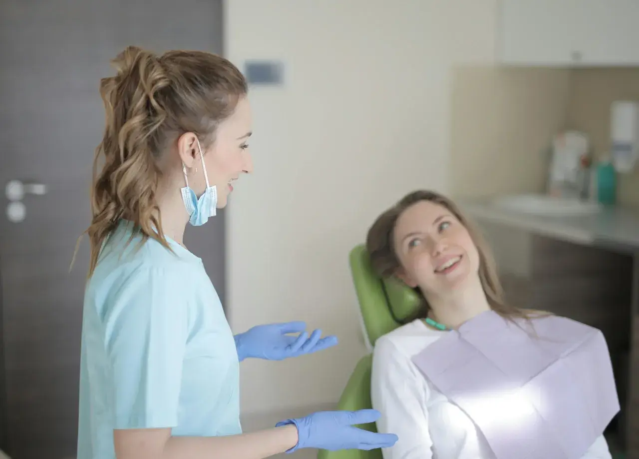 Dentystka w niebieskiej bluzie i rękawiczkach rozmawia z pacjentką. To obrazek pokazujący, jakie są studia stomatologiczne wymagania.
