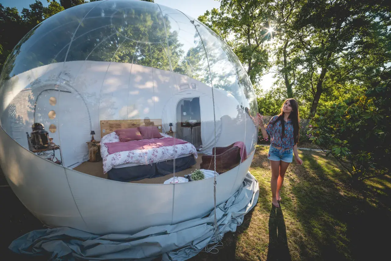 Une jeune femme admire sa **nuit bulle** confortable, un lit douillet dans une sphère transparente au milieu de la nature.