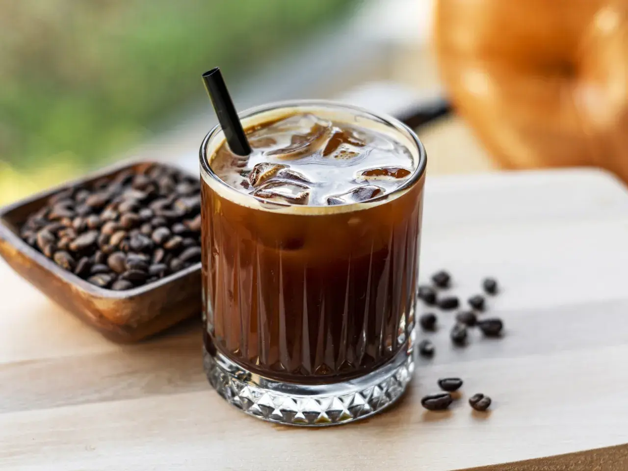 Erfrischender kalter Kaffee mit Eiswürfeln und einem Strohhalm, daneben eine Schale mit Kaffeebohnen.