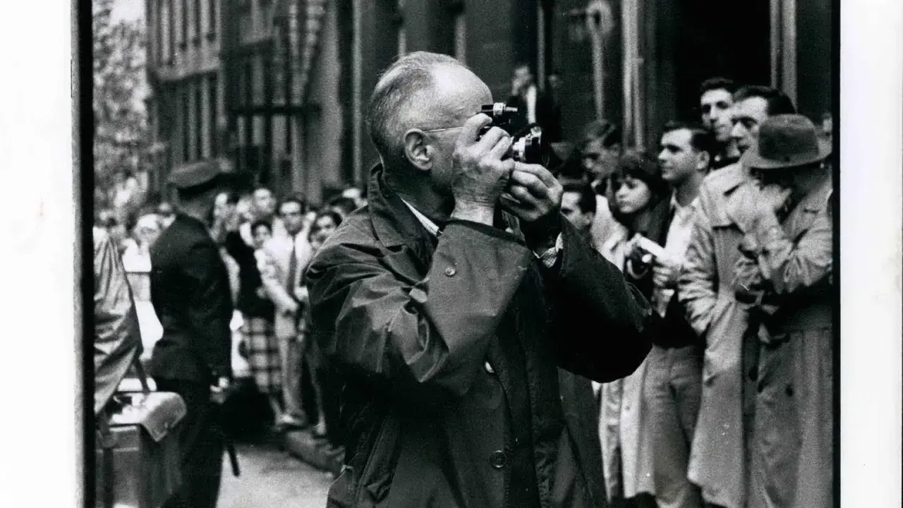Ein Mann mit Brille fotografiert mit einer Leica, umgeben von einer Menge. Ein typisches Bild, das an Henri Cartier-Bresson Fotos erinnert.