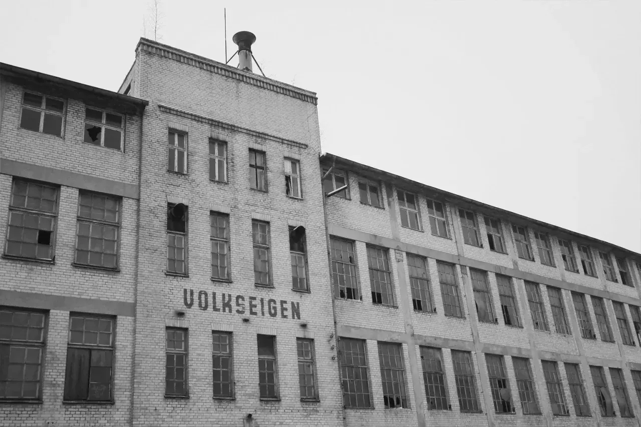 Verlassenes Fabrikgebäude mit der Aufschrift "VOLKSEIGEN", ein Zeugnis der Verstaatlichung in der DDR. Viele Fenster sind zerbrochen.
