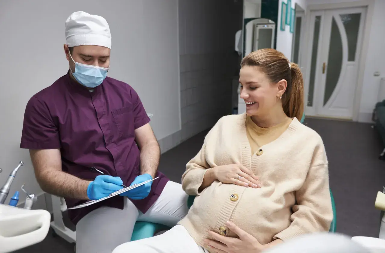 Dentista con mascherina parla con una donna incinta sorridente, discutendo della pulizia dei denti in gravidanza.