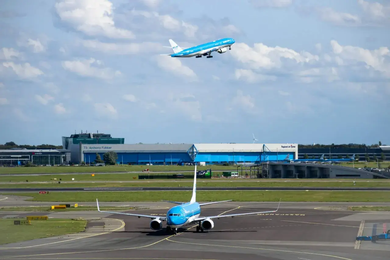 Dwa samoloty linii KLM na płycie lotniska i w powietrzu. Jeden kołuje, drugi wznosi się na tle chmur i budynków lotniska.