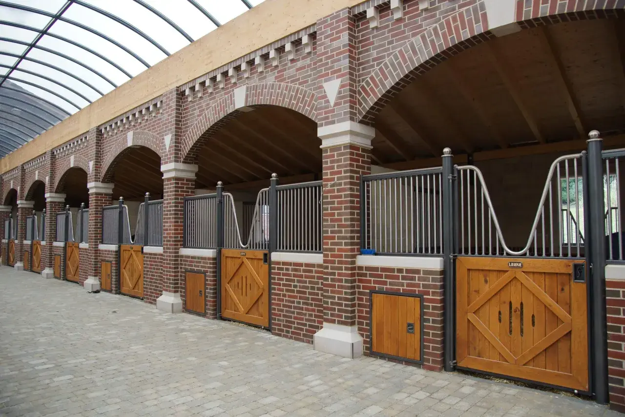 Rangée de stalles de chevaux en briques avec portes en bois et toiture transparente.